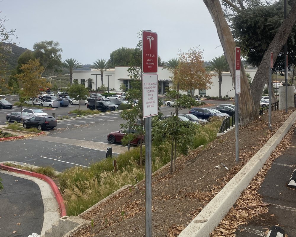 Red Tesla Vehicle Charging Only signs on the slope above the La Costa Supercharger lot on El Camino Real in Carlsbad, the Equinox club across the lot