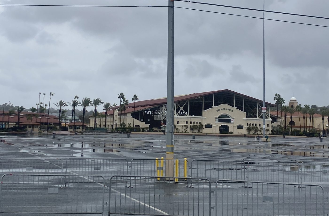 An empty, rain-soaked Del Mar Fairgrounds parking lot lined with crowd barriers, the Del Mar Arena and its long red roof behind it