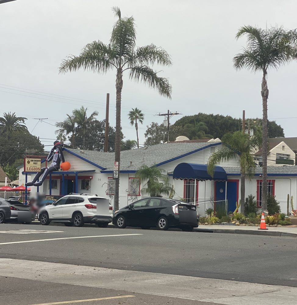 The American Legion San Dieguito Post on F Street in Encinitas under a gray sky, a giant Halloween skeleton on the roof (license plates blurred)