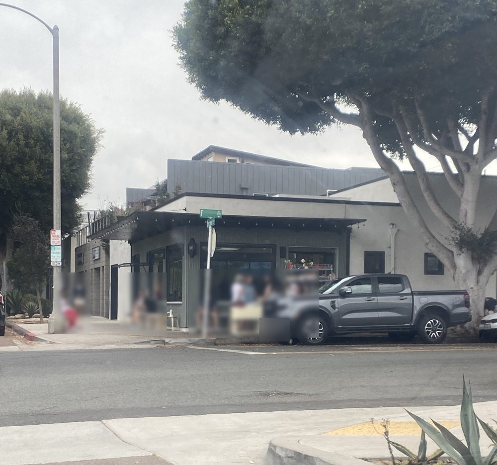 Necessity Coffee's corner storefront at 2nd and F Streets in downtown Encinitas on a gray morning, outdoor pastry rack out front and people waiting (faces blurred)