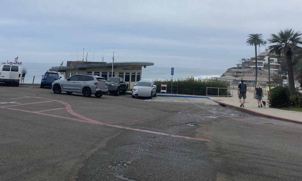 The Moonlight Beach lot and the City of Encinitas Marine Safety Center under a gray sky, whitewater on the water behind it (plates and faces blurred)
