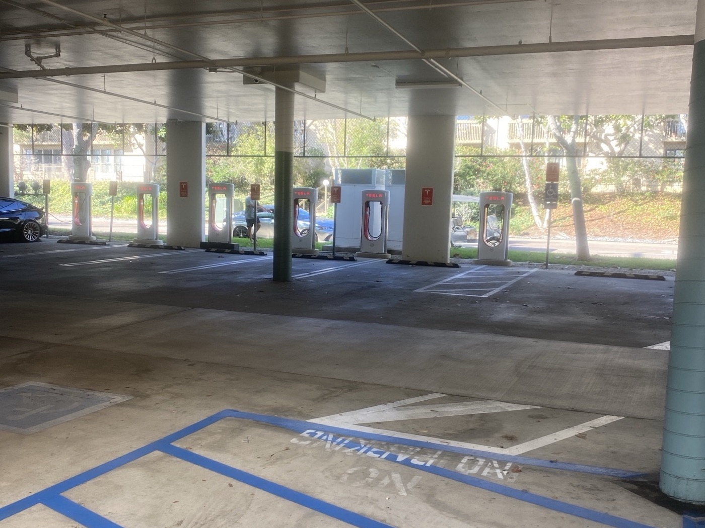 The Solana Beach Supercharger stalls in a covered garage, red Tesla logos lit, one black Tesla charging at the left end, the rest open