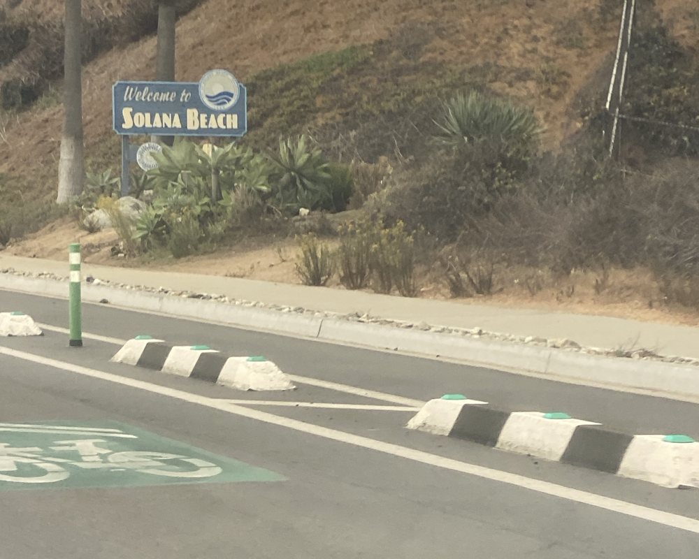 The Welcome to Solana Beach sign on a planted bluff beside Coast Highway 101, a green-painted bike lane with curb separators in front
