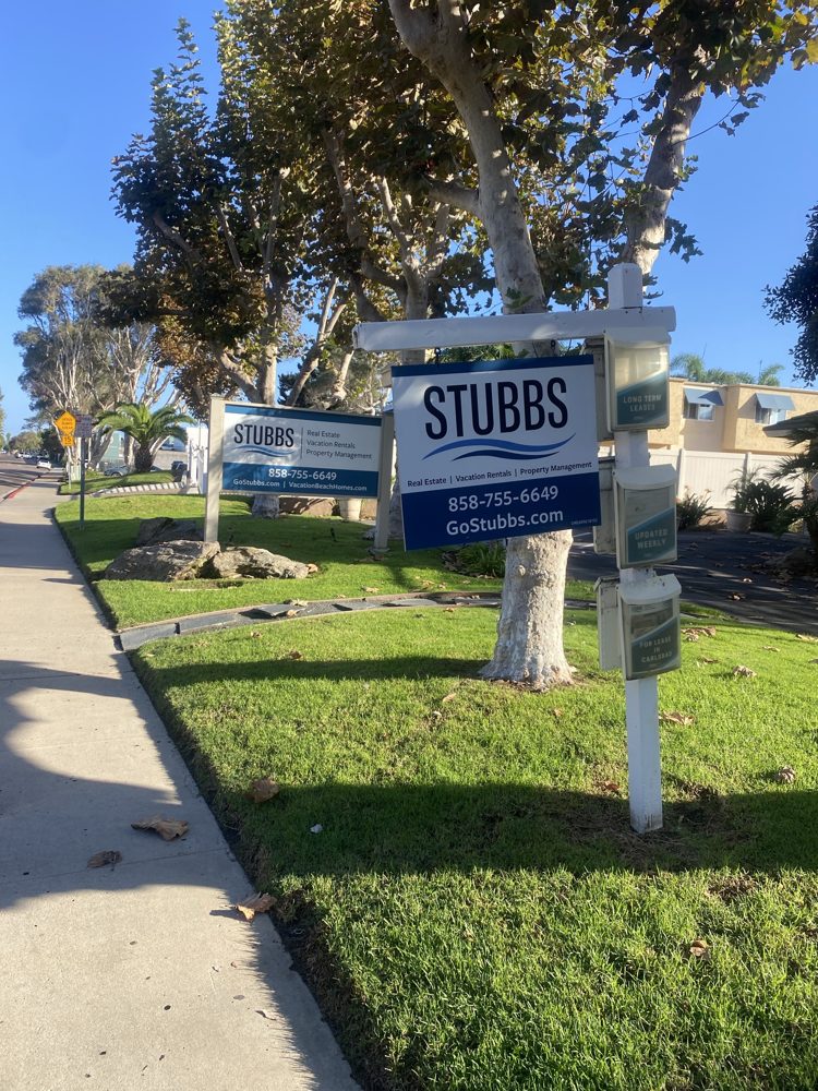 Stubbs signs on a lawn under sycamores on Sierra Avenue: real estate, vacation rentals, property management, with flyer boxes for long-term leases, updated weekly